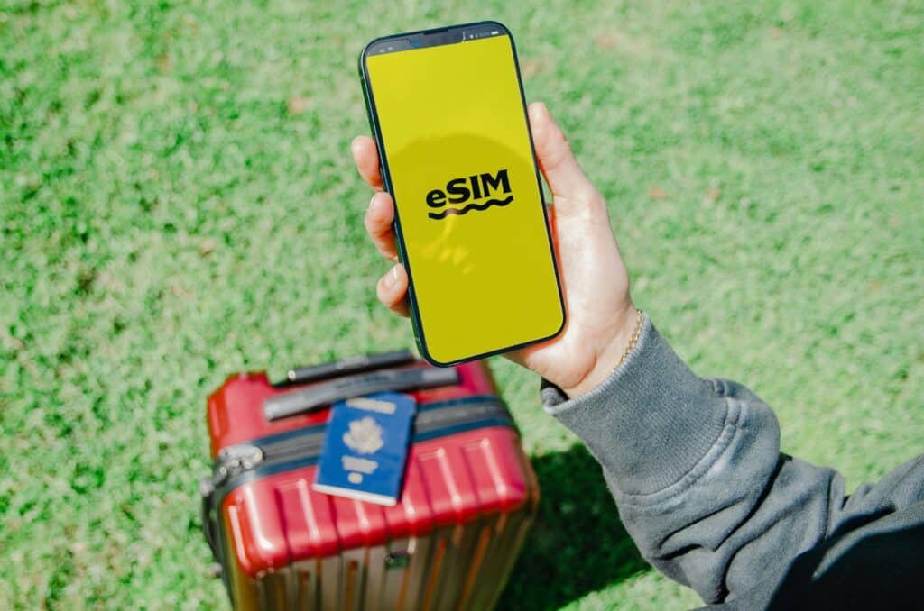 Uma pessoa segura um smartphone com a inscrição "eSIM" em uma tela amarela, acima de uma mala vermelha e um passaporte, na grama verde. Imagem para ilustrar post sobre eSIM Porto Rico.