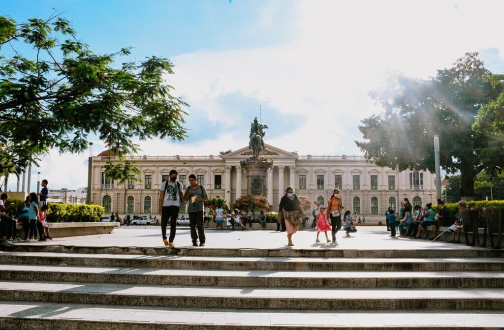 Construção histórica atrás, com pessoa passando à frente e uma pequena escadaria, envolto de natureza nos cantos e ao centro em dia de céu azul e ensolarado. 