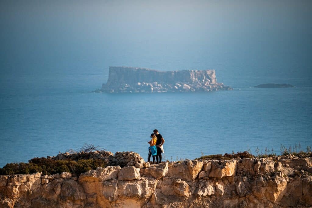 Homem e mulher em um Mirante de pedras, olhando algo que está nas mãos da menina, com vista do mar e de uma pequena ilha no meio, num dia ensolarado