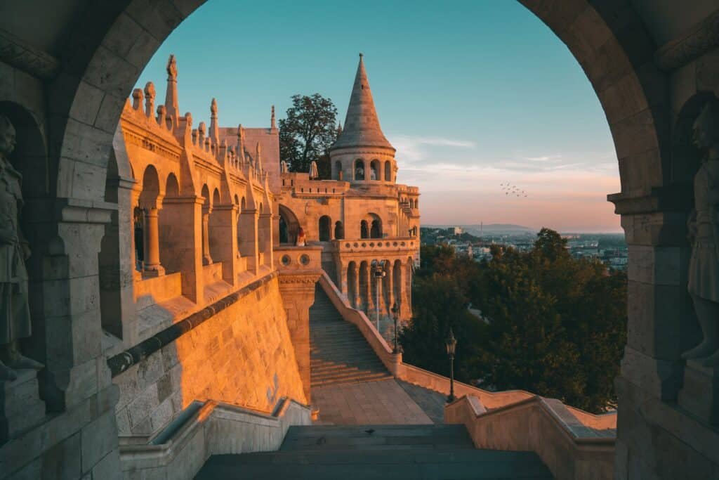 Um arco emoldura a foto de um castelo bege para ilustrar o post sobre seguro viagem Hungria. Há algumas árvores ao redor e uma mulher explorando o local. - Foto: Tobias Reich via Unsplash